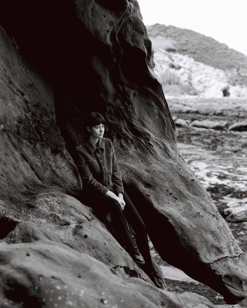 A woman sits on a large rock formation with a rocky landscape and hill in the background.