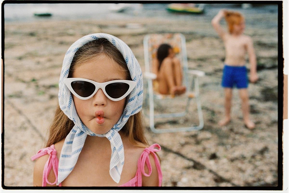 A girl wearing a headscarf and sunglasses makes a funny face at the beach.