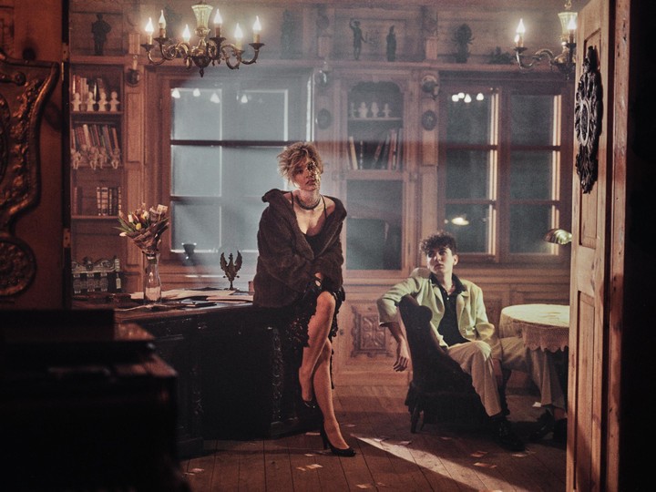 A woman in a fur coat sits on a desk while a man sits in a chair in a room with chandeliers and bookshelves.