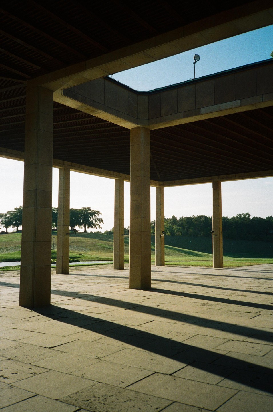 Sunlight casts long shadows from concrete pillars onto a paved ground, with a grassy hill and trees in the background.