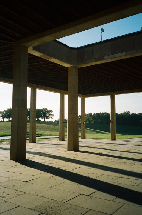 Sunlight casts long shadows from concrete pillars onto a paved ground, with a grassy hill and trees in the background.