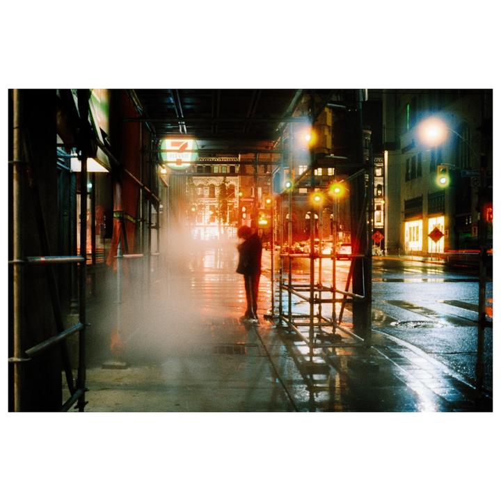 A person stands on a wet city street at night, surrounded by scaffolding and illuminated by streetlights and building lights.