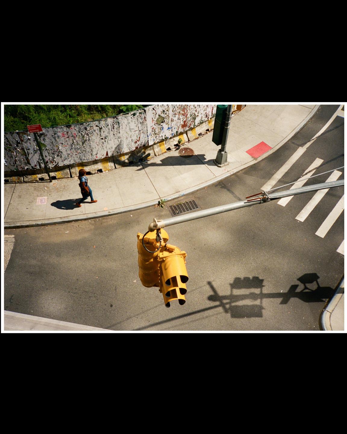 A woman walks on a sidewalk next to a street with a yellow traffic light hanging overhead.