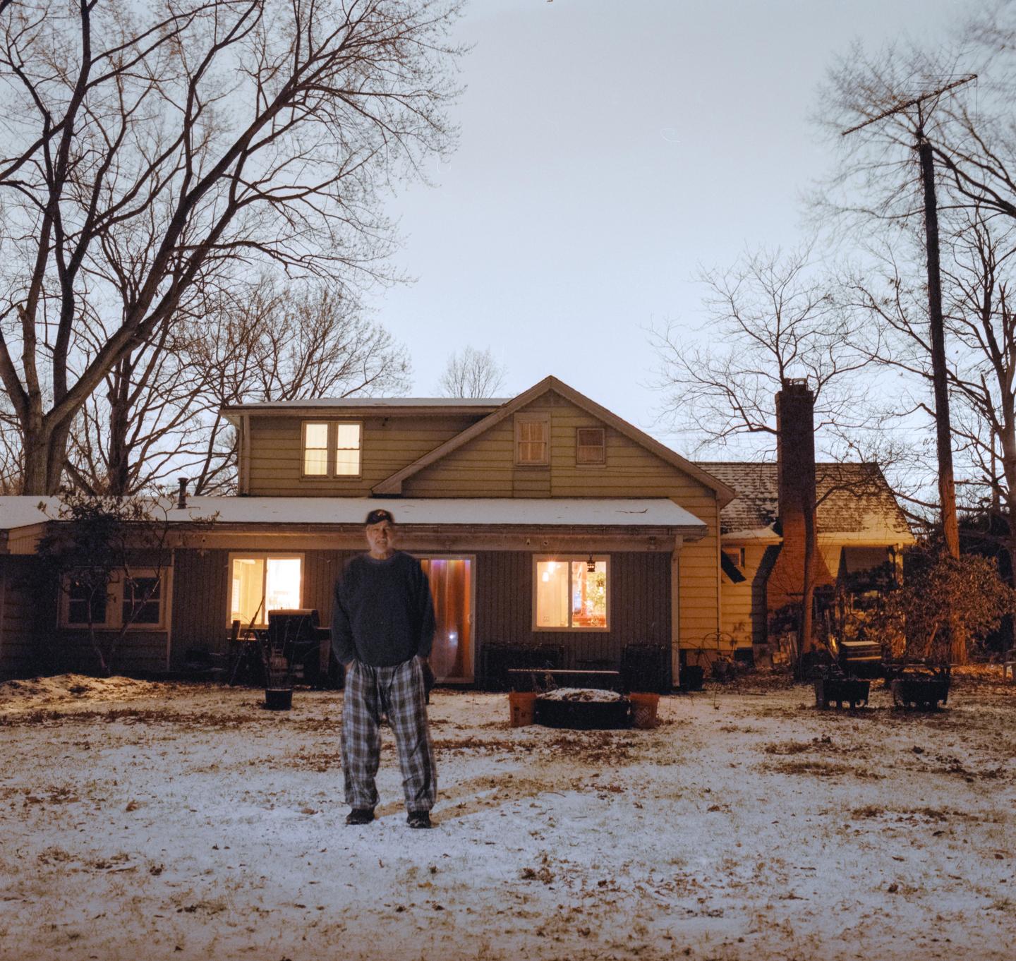 A man stands in a snowy yard in front of a house with illuminated windows at dusk.