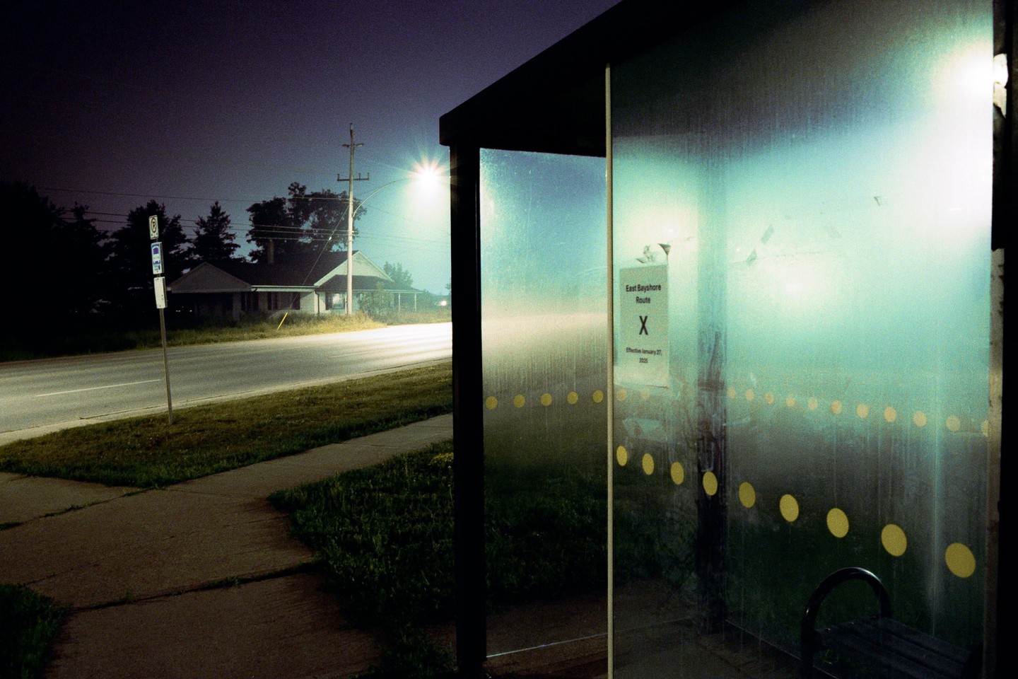 A bus stop shelter with a sign for "East Bayshore Route X" stands beside a road at night, with a house visible in the distance under a street light.