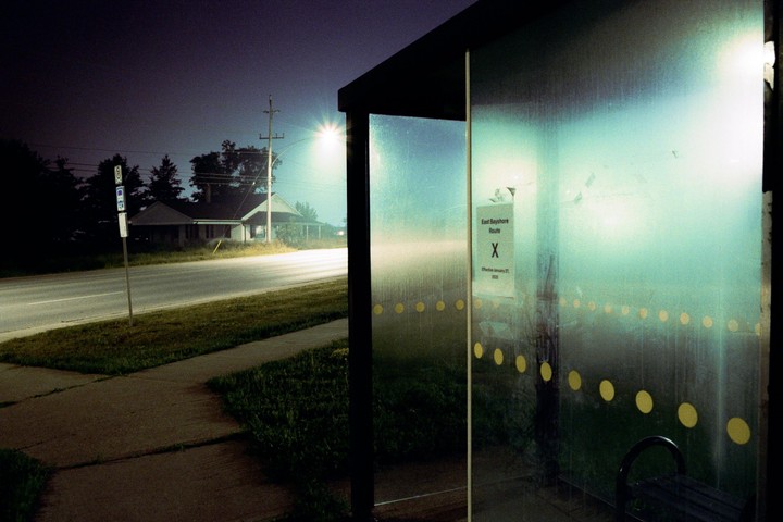 A bus stop shelter with a sign for "East Bayshore Route X" stands beside a road at night, with a house visible in the distance under a street light.