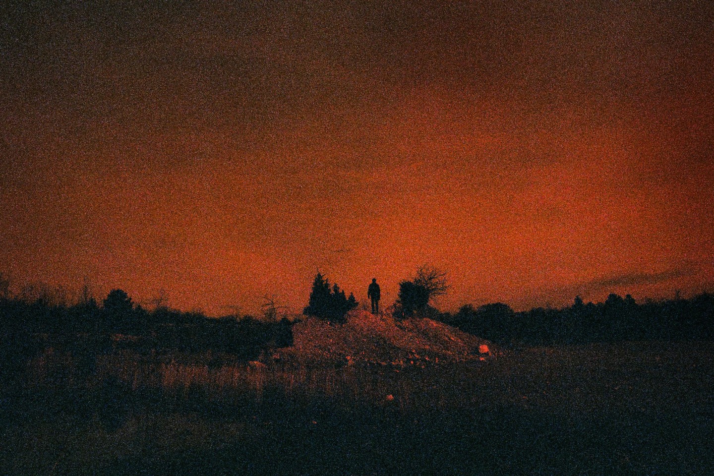 A lone man stands atop a hill silhouetted against a vibrant red sky.