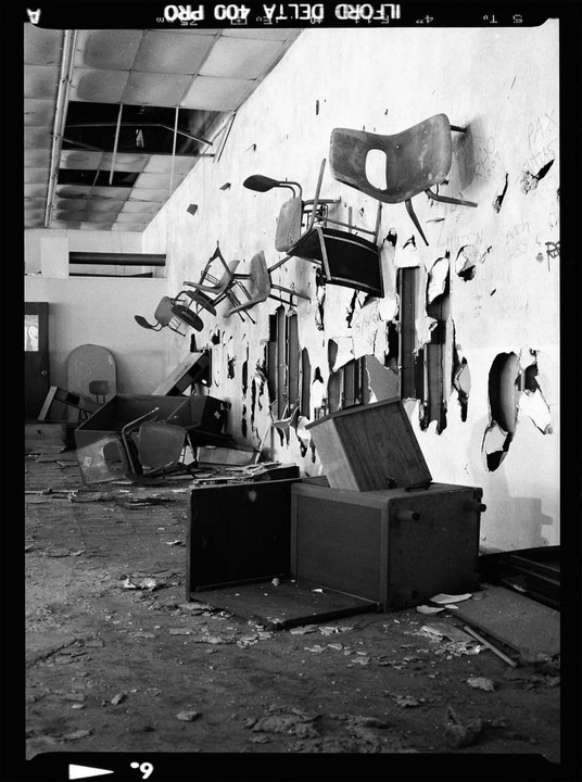 Chairs are mounted on a damaged wall in a derelict room with debris on the floor.