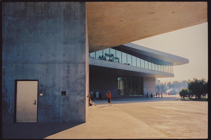 A modern concrete building with large windows stands next to a plaza where people are gathered.