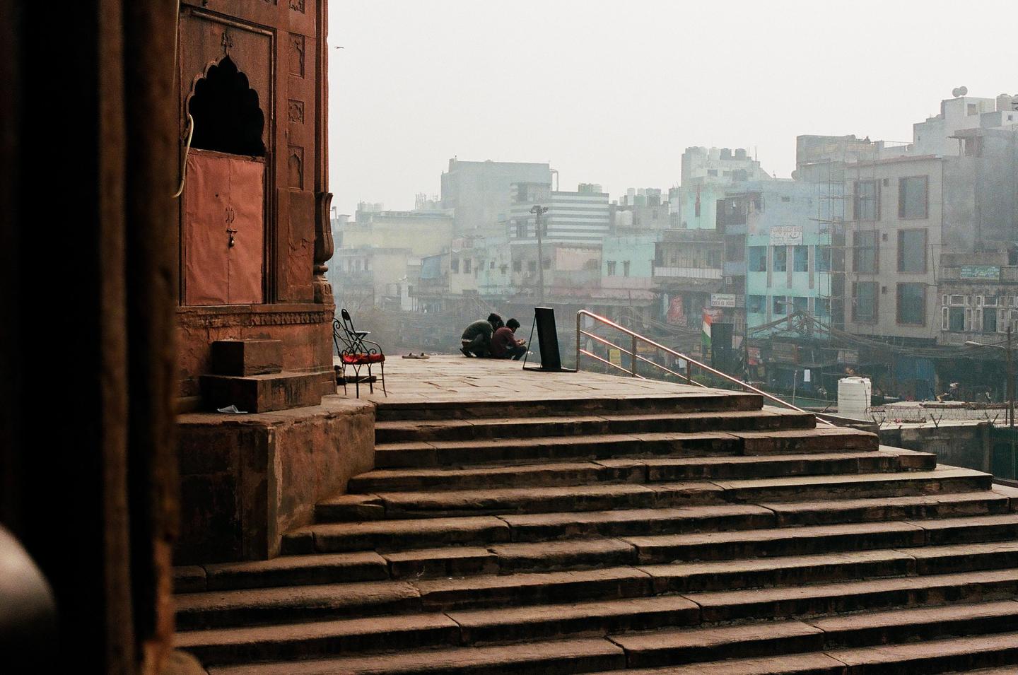 Three men are sitting on a stone platform with a city in the background, viewed from a stone staircase.