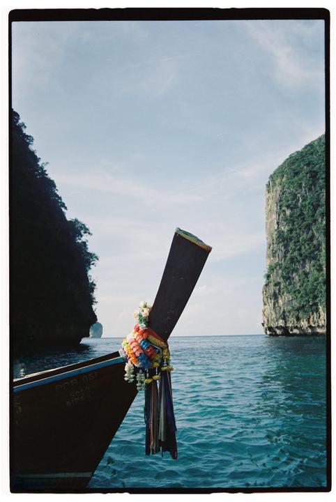 A boat with colorful garlands on its bow sails through turquoise water past two large, rocky islands covered in lush green foliage.