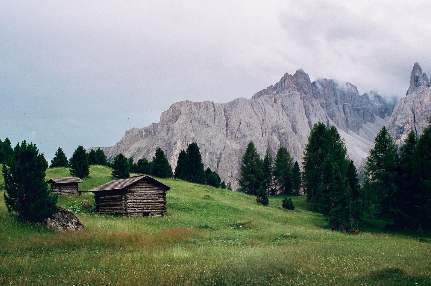 Two wooden cabins sit on a grassy hill with pine trees, overlooking a large mountain range under a cloudy sky.
