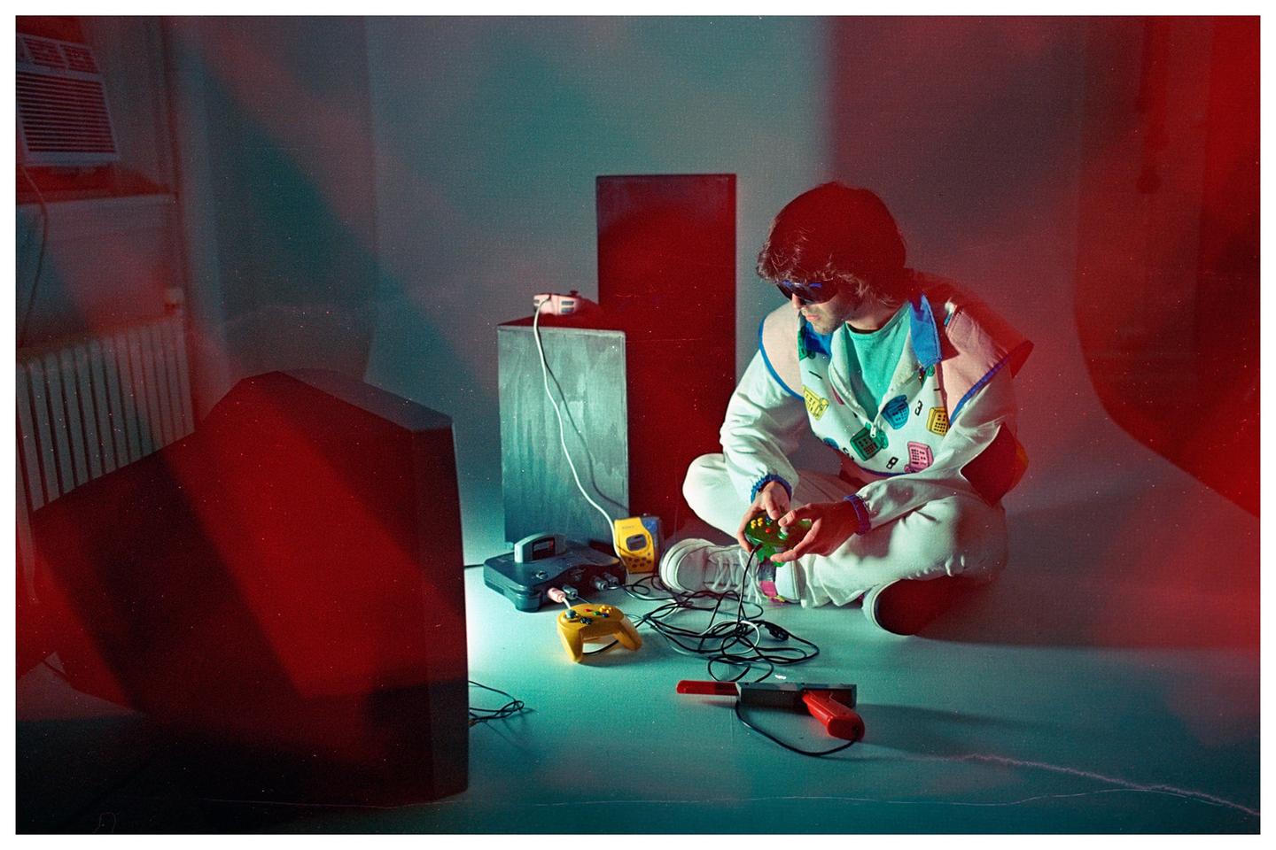 A man in sunglasses and a colorful jacket sits on the floor playing a video game with a console and television in front of him.