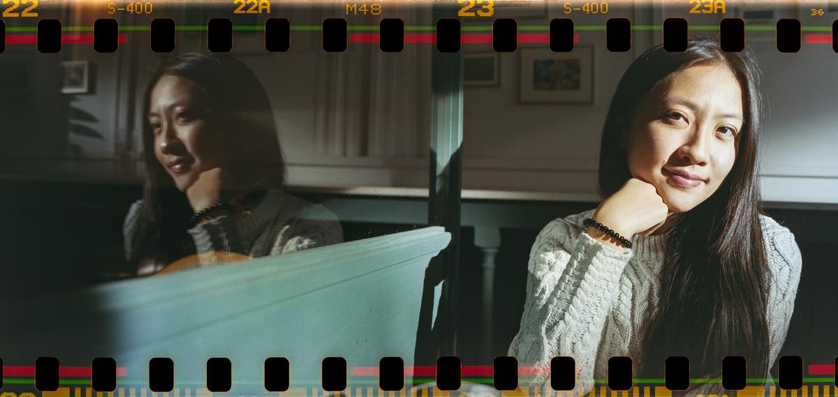 A woman with long hair and a light-colored sweater sits with her chin resting on her hand, with her reflection visible in a surface to her left.