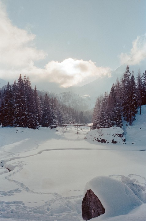 A snow-covered landscape features a wooden bridge over a frozen river, surrounded by pine trees and mountains under a cloudy sky.