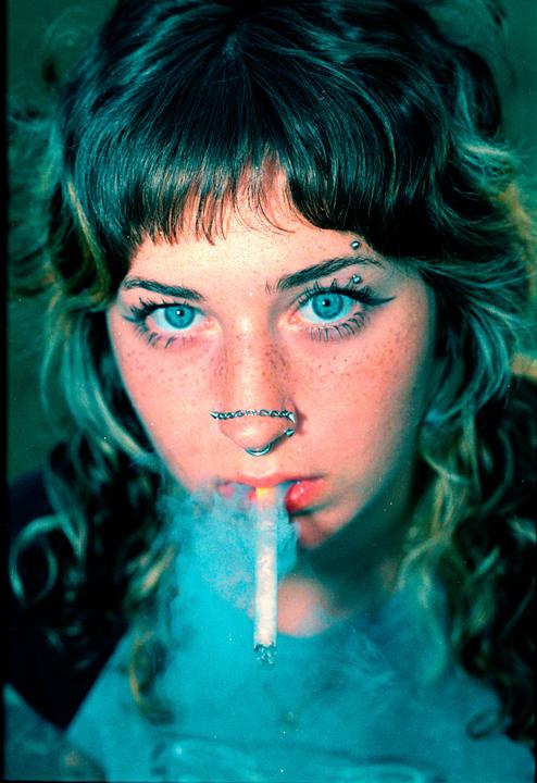 A young woman with blue eyes and freckles exhales smoke from a cigarette.