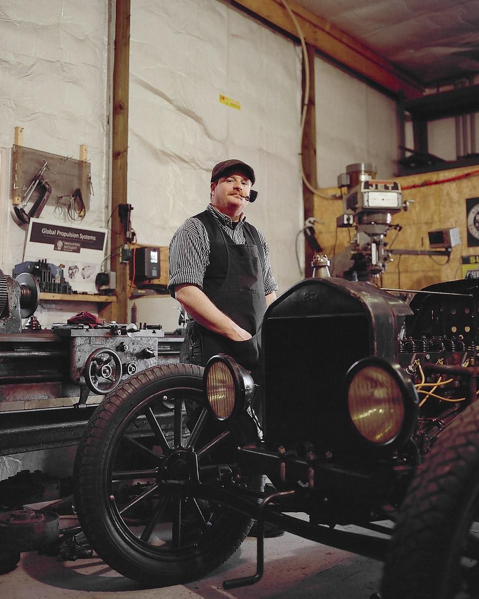 A man with a pipe in his mouth stands next to a vintage car in a workshop.