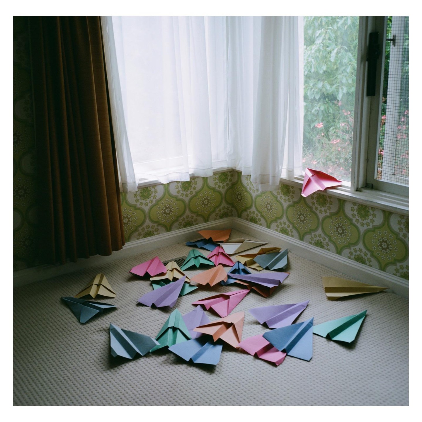 Many colorful paper airplanes are scattered on a carpeted floor next to a window with sheer white curtains and patterned wallpaper.