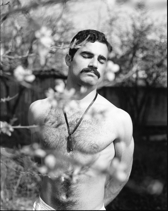 A shirtless man with a mustache and closed eyes stands among blurred foliage.