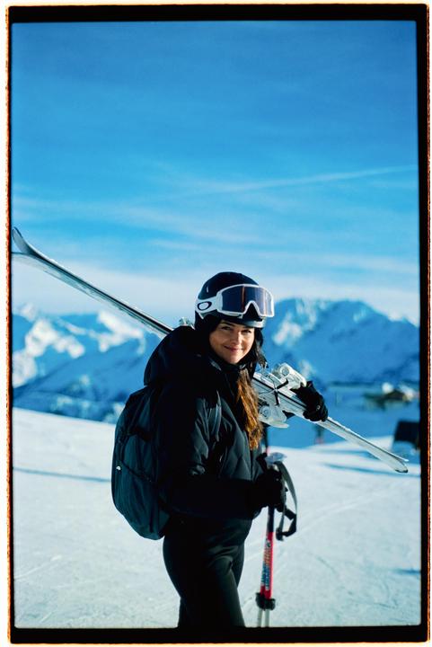 A woman wearing ski gear and carrying skis and poles stands on a snowy mountain with a clear blue sky.