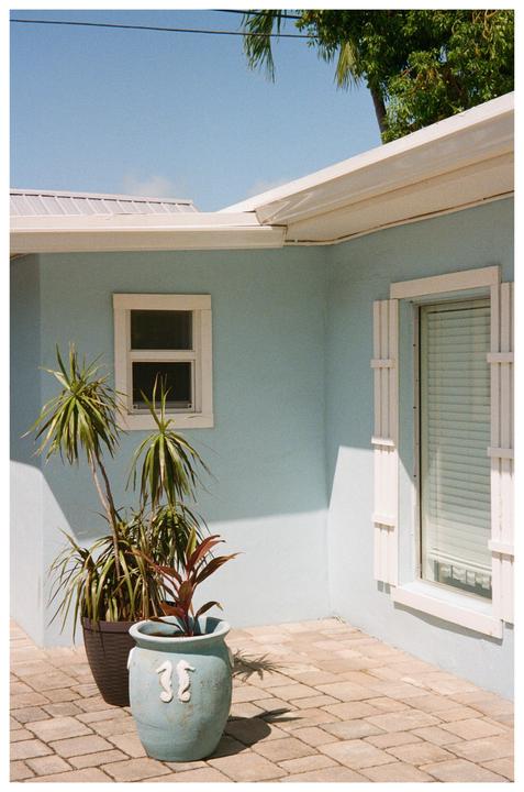 A light blue house with white trim and two potted plants sits on a paved patio under a clear sky.