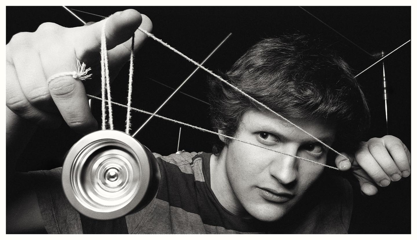 A man with a serious expression looks at the viewer while holding a yo-yo with strings crisscrossing in front of his face.