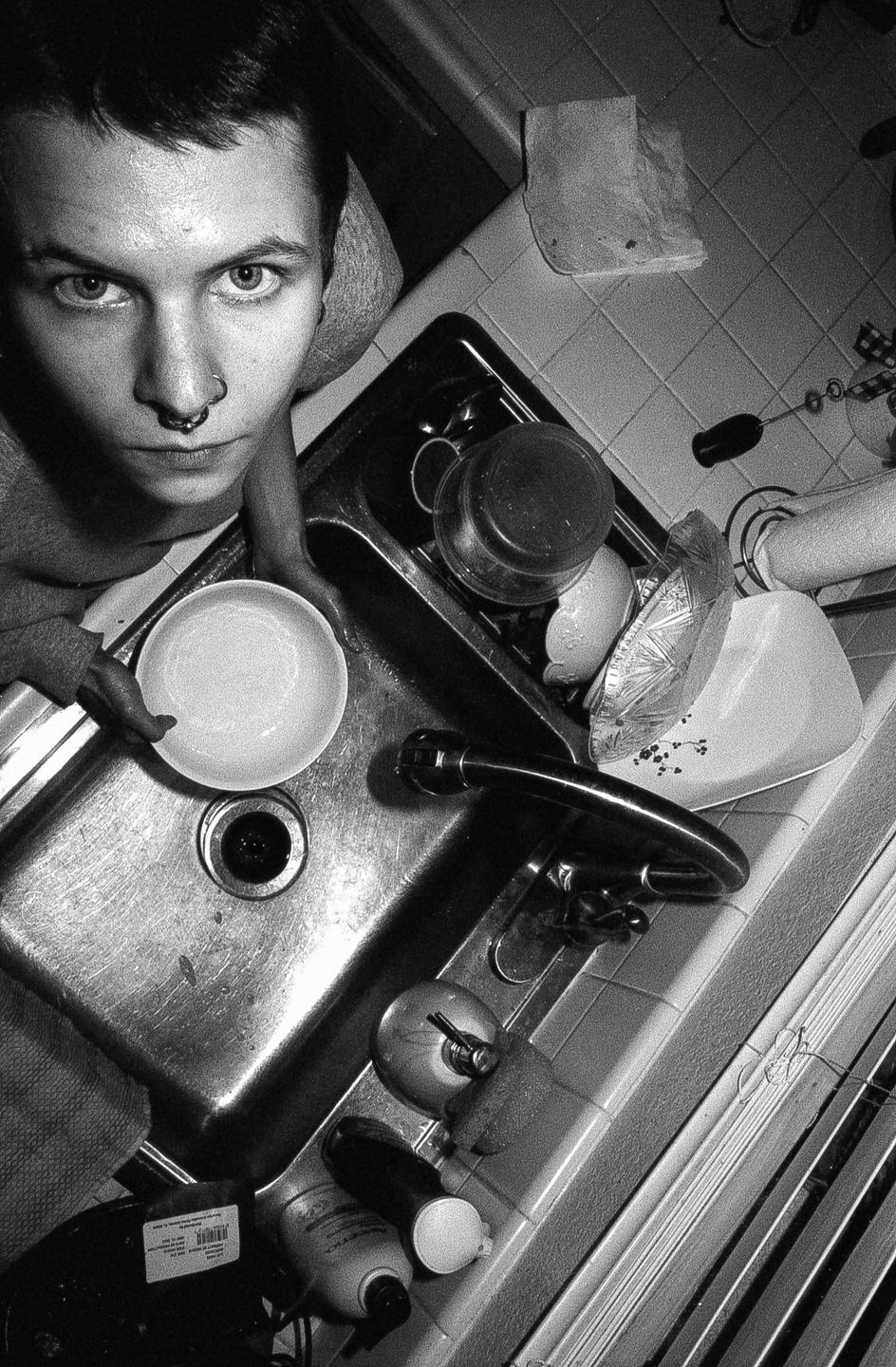 A man with a nose ring looks up at the camera while holding a bowl over a kitchen sink full of dishes.