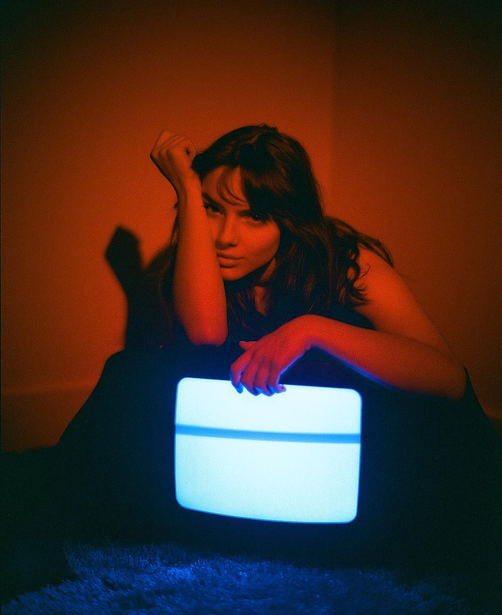A woman sits on the floor with her arm resting on a glowing television screen.