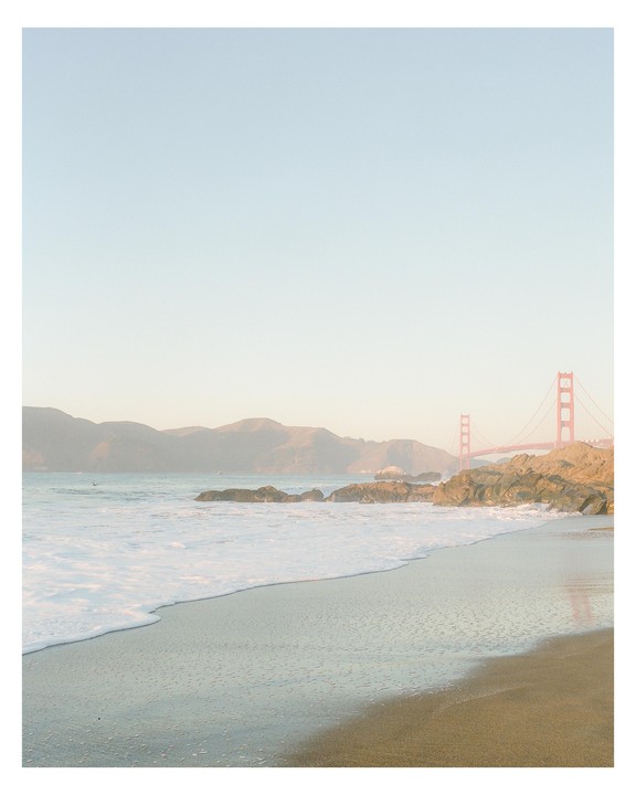 Waves crash on a sandy beach with a bridge and hills in the background.