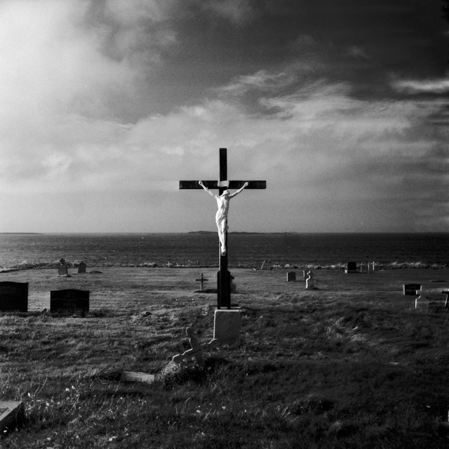 A crucifix stands in a grassy cemetery overlooking the ocean under a cloudy sky.