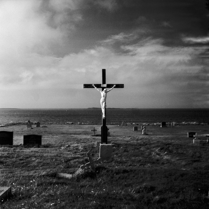 A crucifix stands in a grassy cemetery overlooking the ocean under a cloudy sky.