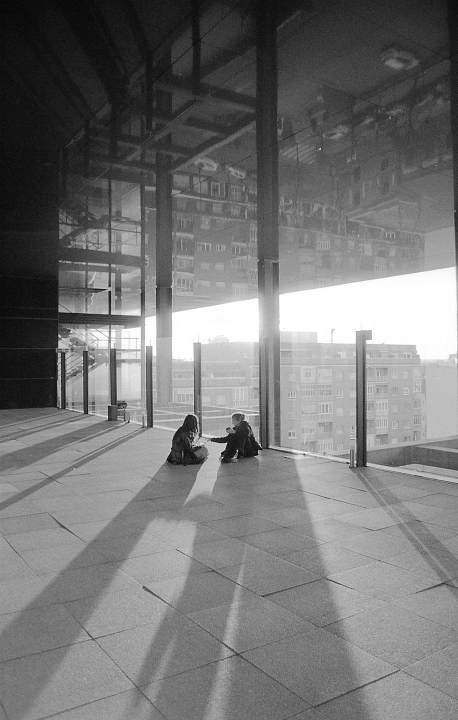 Two people sit on a tiled floor next to a large window with a city visible outside.