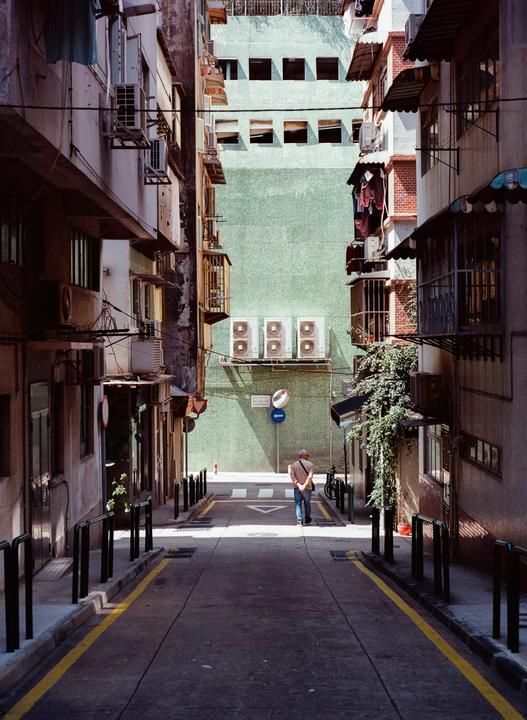 A man walks down a narrow street lined with tall buildings towards a light green wall with air conditioning units.