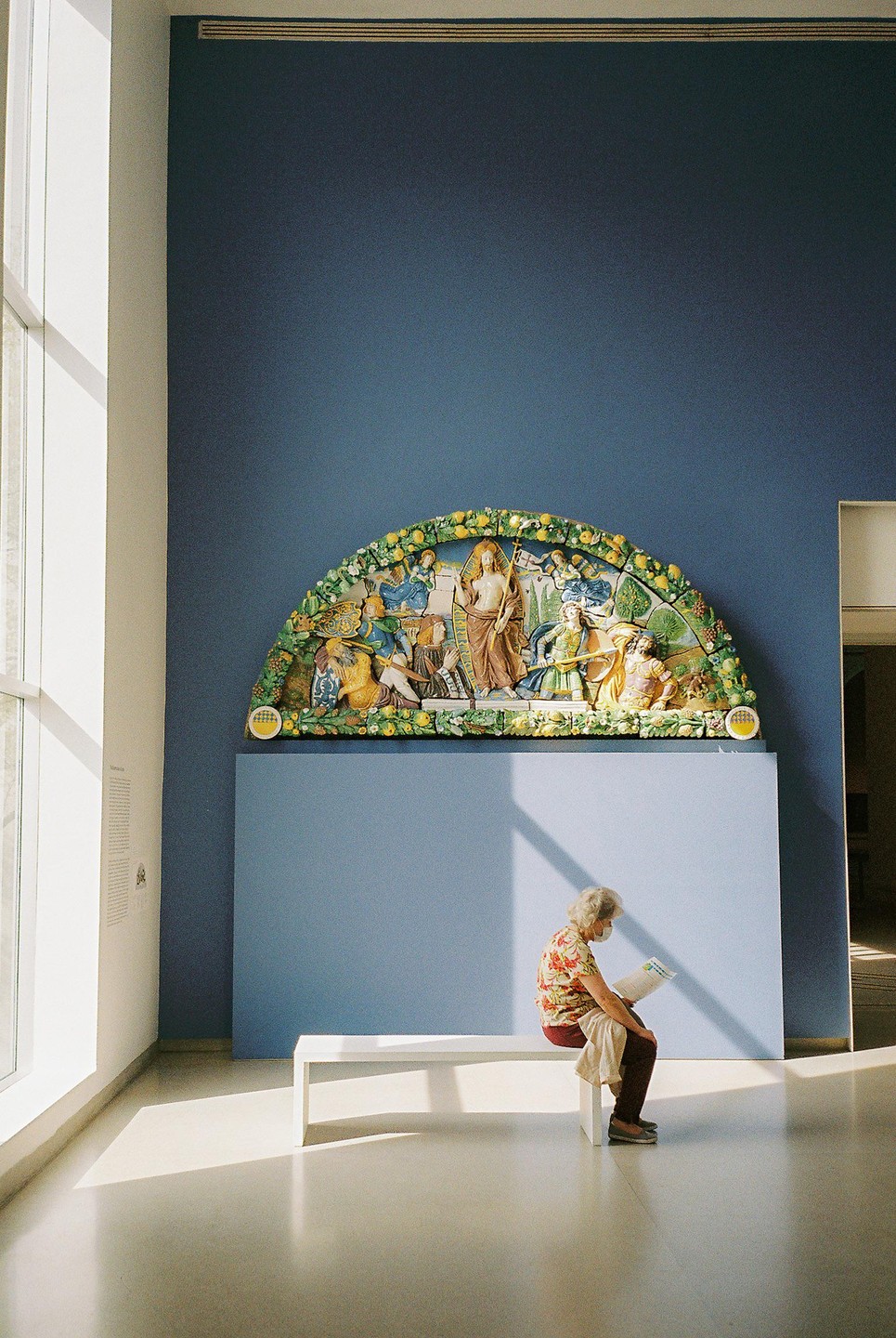 A woman sits on a bench in a museum gallery, reading a pamphlet, with a large colorful relief sculpture on the wall behind her.