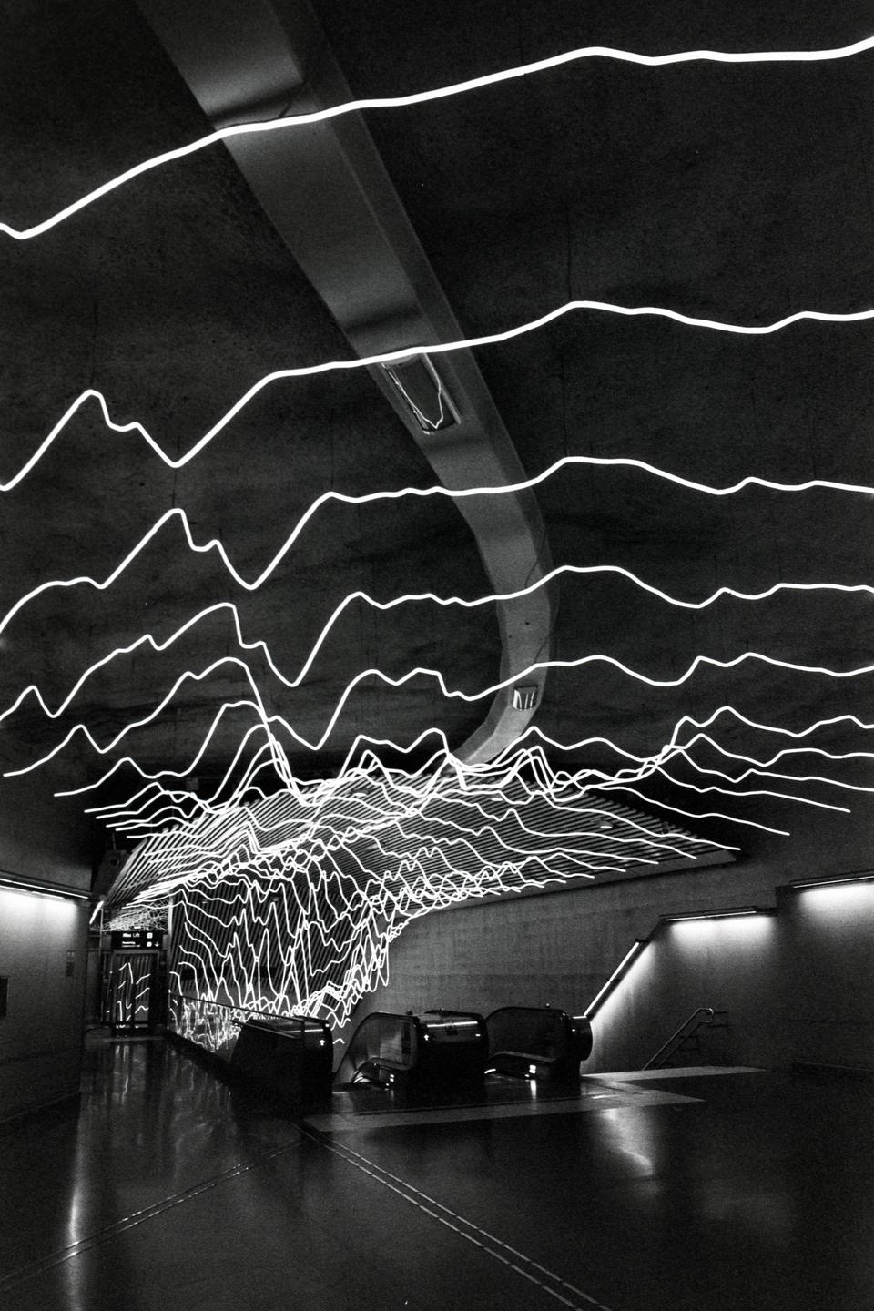 A subway station features a ceiling installation of wavy neon lights that resemble a sound wave.