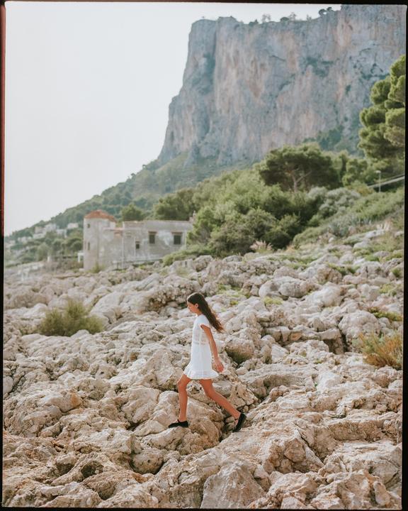 A girl in a white dress walks across a rocky landscape with a large cliff and buildings in the background.