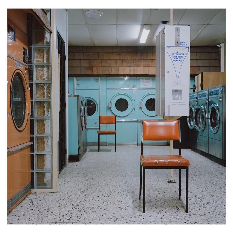 Two orange chairs sit on a speckled floor in a laundromat with rows of washing machines.