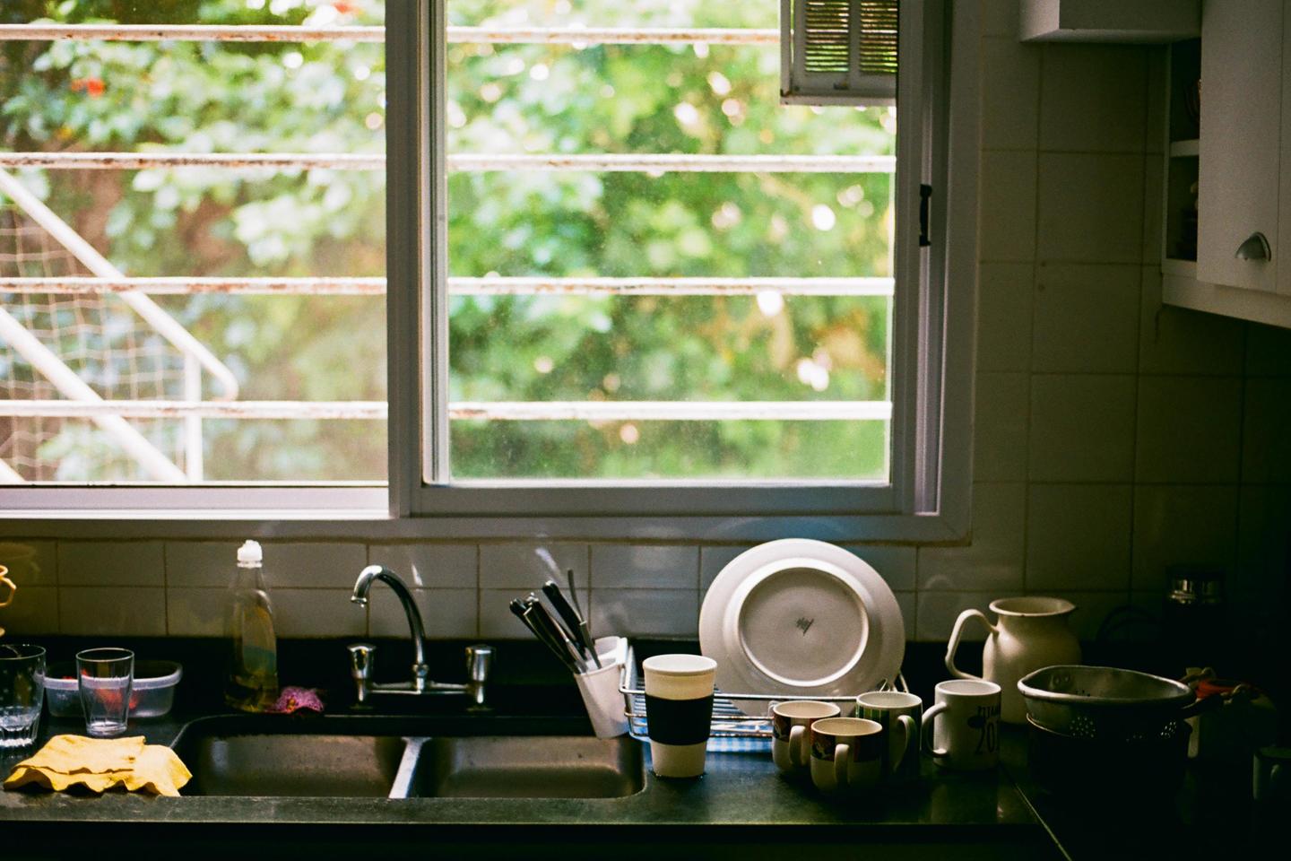 A kitchen sink and counter are filled with dishes and utensils next to a window looking out onto green foliage.