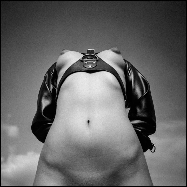A woman's torso is visible from below, wearing a leather harness and jacket against a cloudy sky.