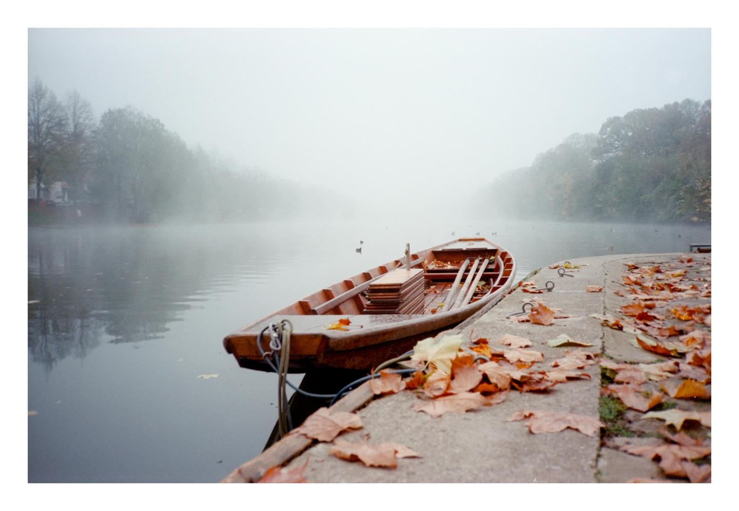 A wooden rowboat is moored to a concrete bank covered in autumn leaves, with a foggy lake and trees in the background.