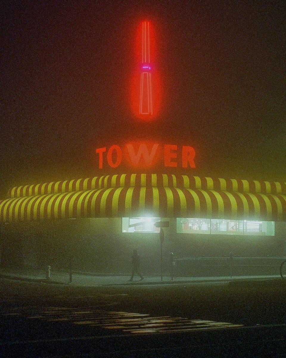 A person walks past a building with a striped awning and a neon sign that reads "TOWER" in the fog at night.