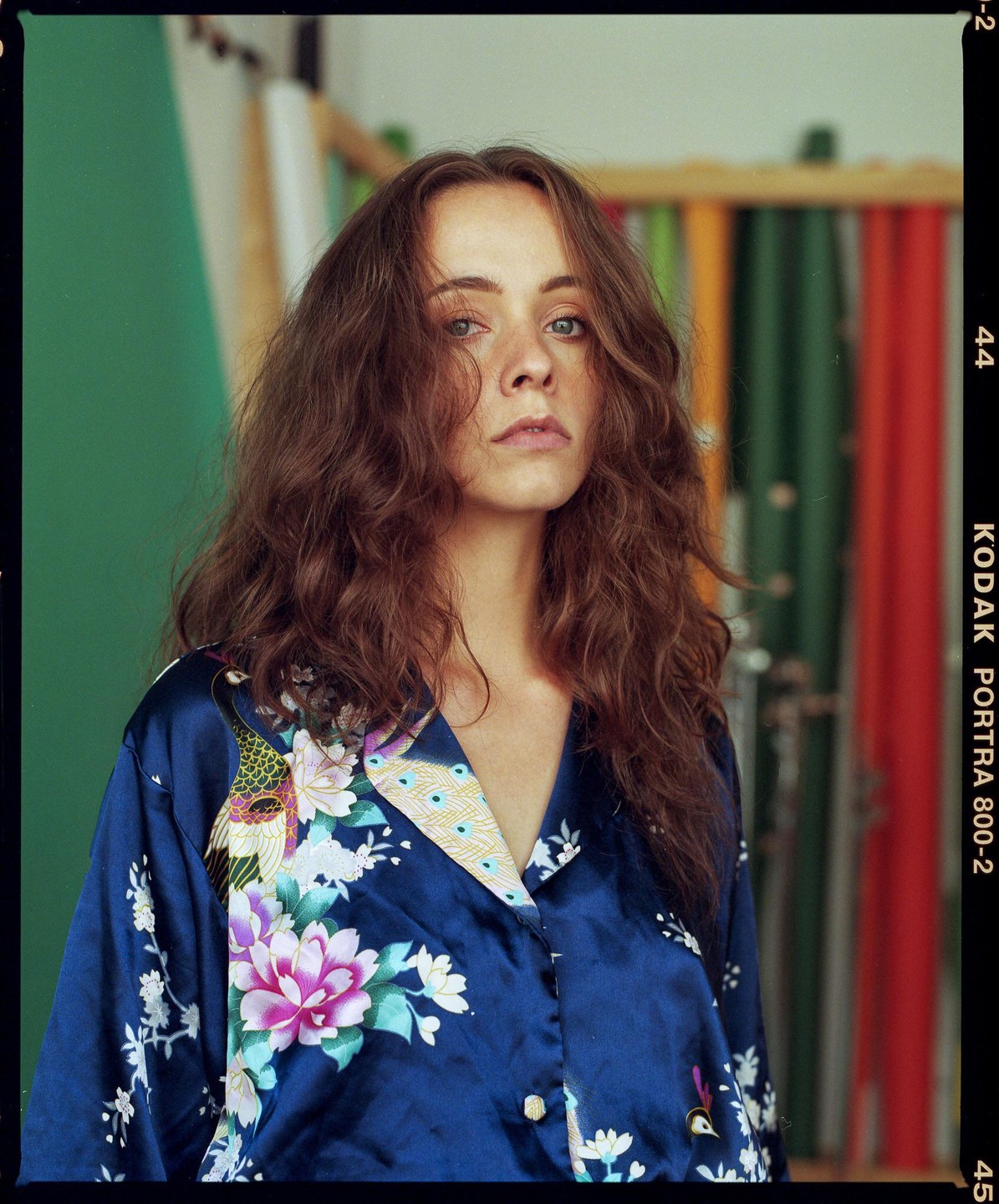A woman with long, wavy hair wears a blue silk shirt with a floral and peacock pattern.