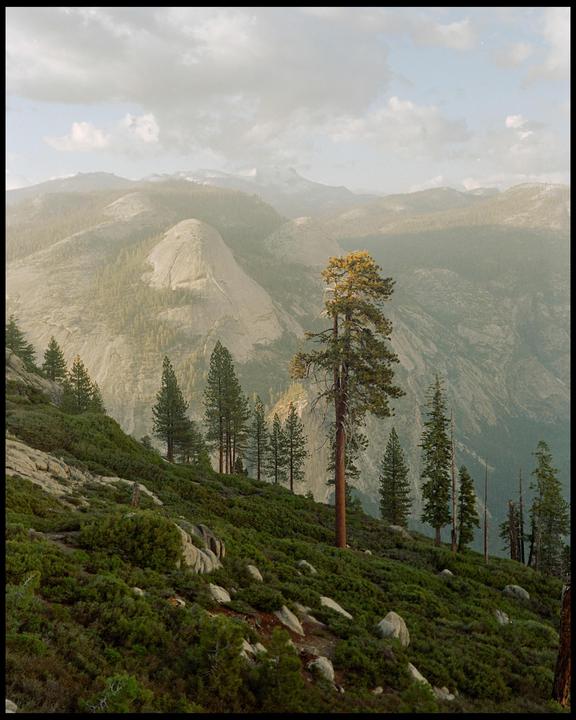 A mountain landscape shows a hillside covered in green foliage and trees, with a large rock formation and more mountains in the background under a cloudy sky.