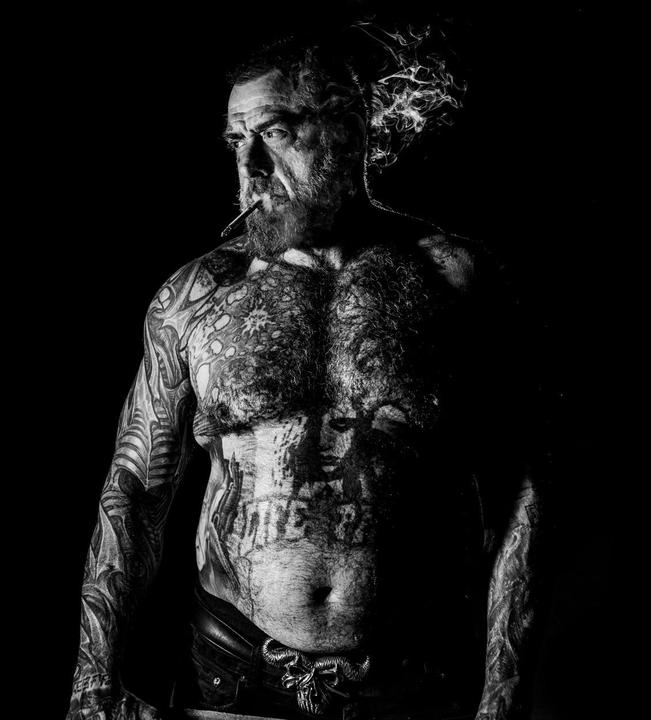 A shirtless man with a beard and tattoos smokes a cigarette, with smoke rising from his mouth.
