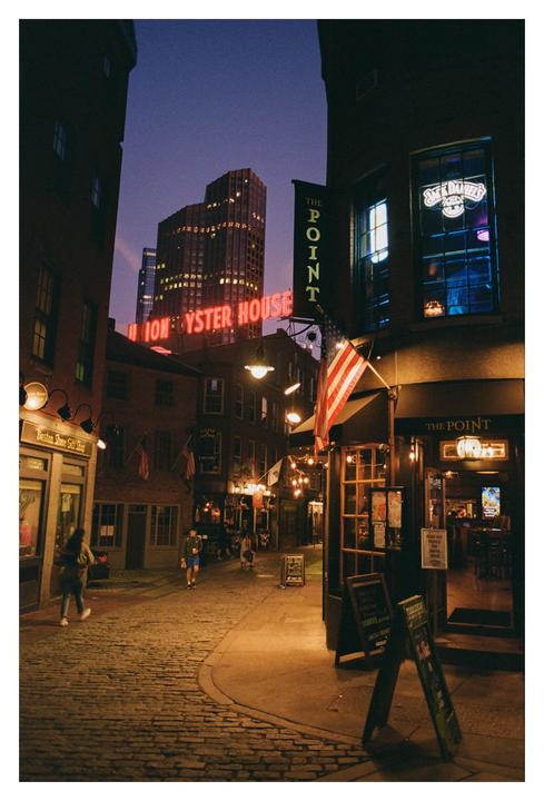 A cobblestone street is illuminated by neon signs and string lights at night, with people walking along the street.