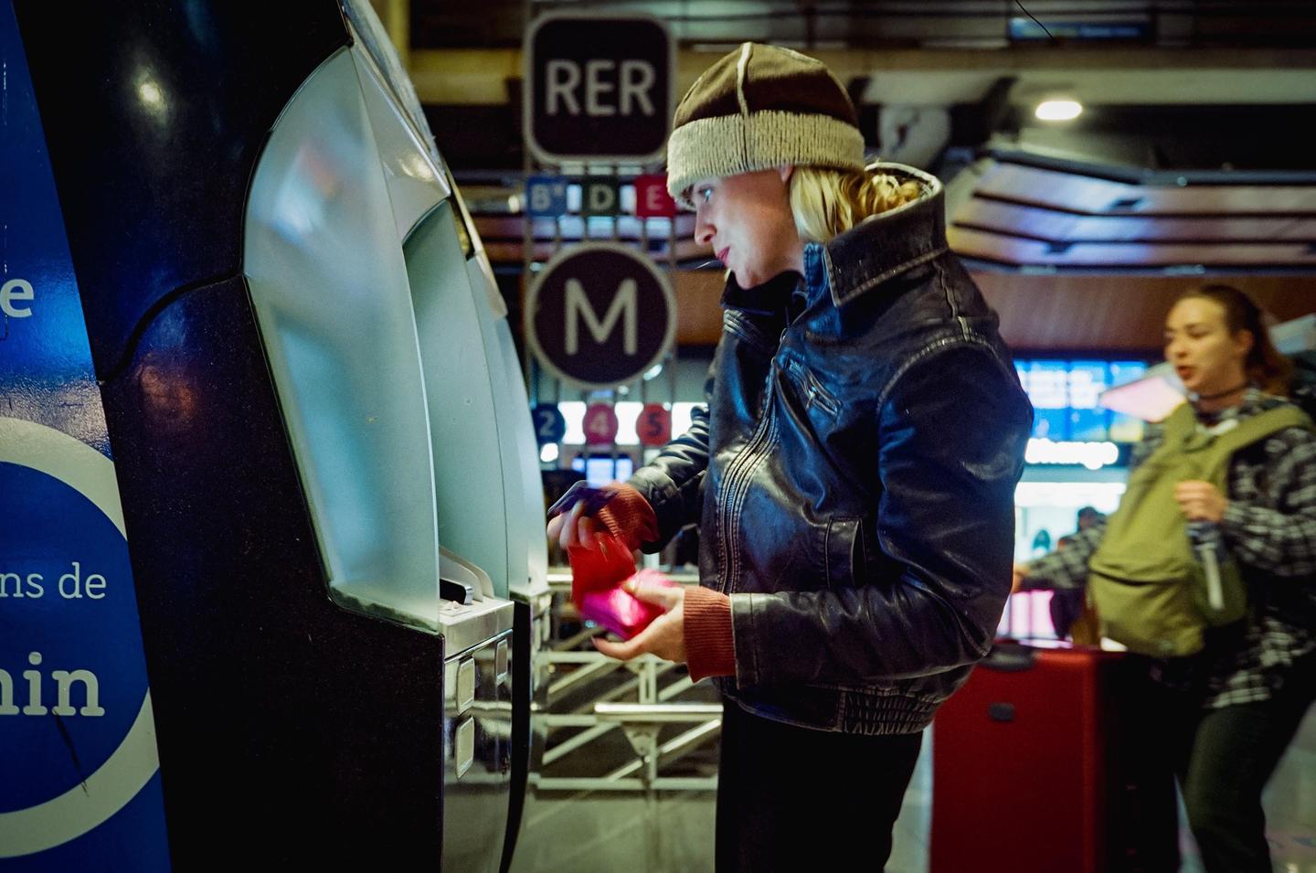 A woman in a leather jacket and knit hat uses a ticket machine in the Paris Metro.
