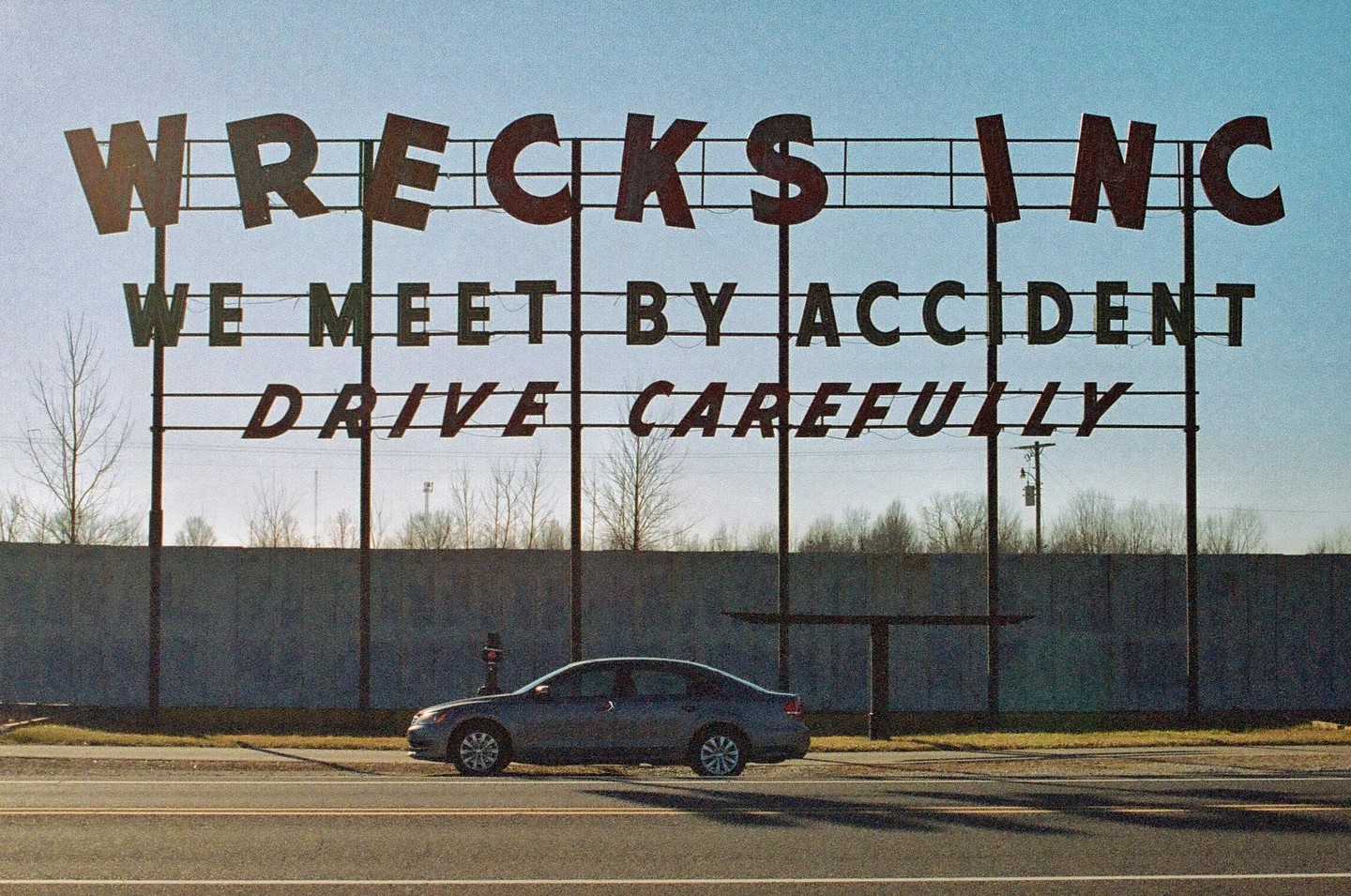 A car is parked in front of a large sign that reads "WRECKS INC WE MEET BY ACCIDENT DRIVE CAREFULLY" under a clear sky.