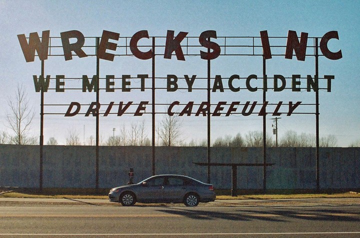 A car is parked in front of a large sign that reads "WRECKS INC WE MEET BY ACCIDENT DRIVE CAREFULLY" under a clear sky.