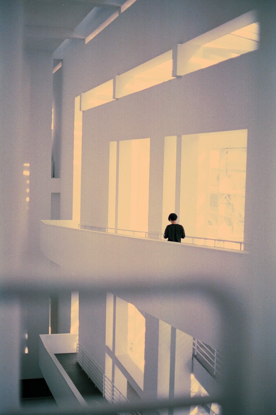 A person stands on a balcony overlooking a multi-story building interior with strong light and shadow patterns.