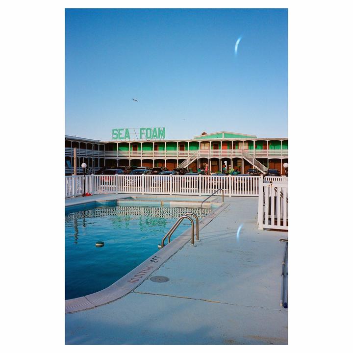 A swimming pool with a white fence in front of a motel with a "SEA FOAM" sign is visible under a clear sky.